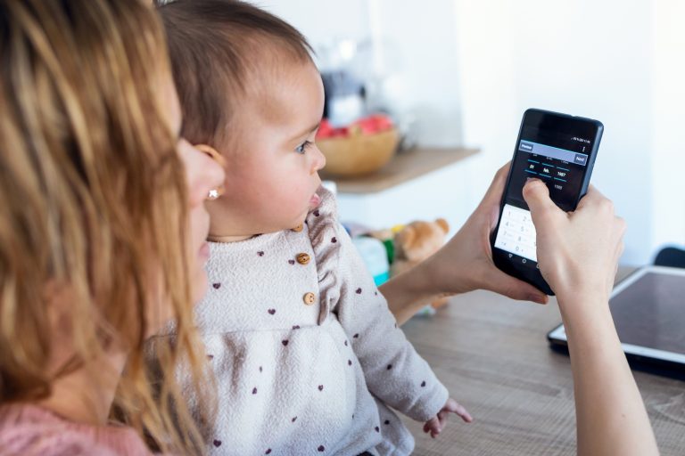 Young woman using her mobile phone while her baby girl looks at it at home.